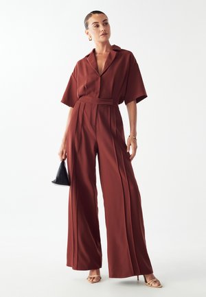 Einteiler in Rostfarbe mit klassischem Kragen, kurzen Ärmeln, weitem Bein und tailliertem Design. Glatte Stoffqualität mit Falten.