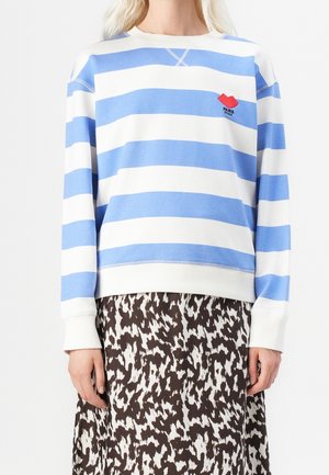 Vrouw die een blauw-wit gestreepte sweatshirt draagt met een rode lippenafdruk en de tekst "PARIS", gecombineerd met een zwart-witte geruite rok.
