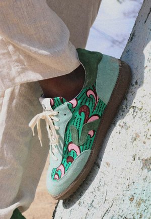 Pied portant une basket verte et rose à motifs avec des lacets blancs, reposant sur une surface blanche texturée, pantalon beige visible.