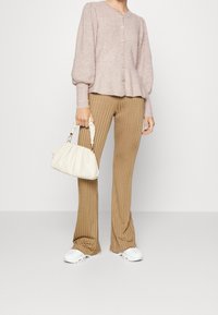 Mujer vistiendo pantalones beige con relieve, suéter abotonado en color malva claro, zapatillas blancas, sosteniendo un pequeño bolso de marfil con asa fruncida.