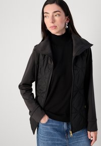 Een vrouw met lang, donker haar draagt een zwarte ritsjack over een trui, terwijl ze tegen een lichtgrijze achtergrond staat, met haar handen in de zakken.