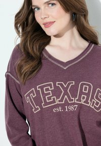 Maglietta a maniche lunghe color bordeaux con scollatura a V, decorata con la scritta "TEXAS" e "est. 1987" in lettere beige, accentuata da cuciture a contrasto.