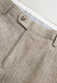 Pantalon beige à fines rayures avec passants pour ceinture, fermeture avant par boutons, et doublure intérieure grise à carreaux partiellement visible.