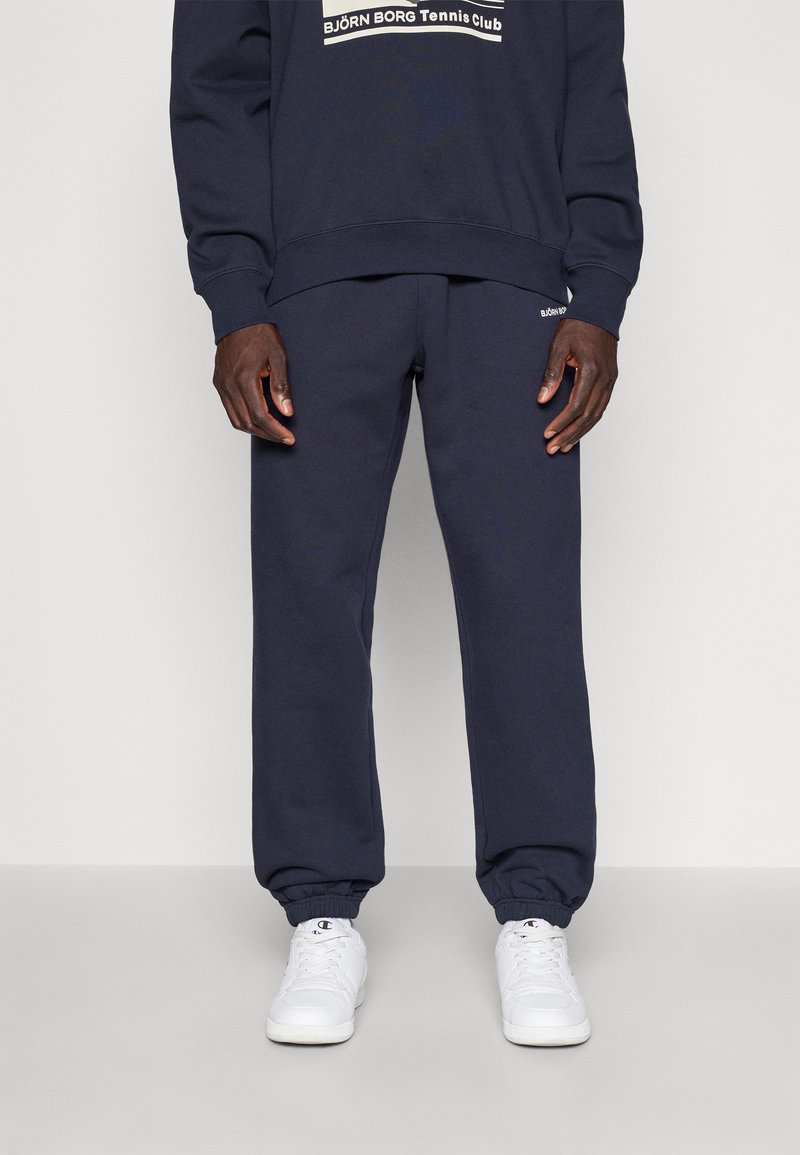 Σκούφος μπλε φούτερ και αντίστοιχες φόρμες, φτιαγμένα από μαλακό υλικό. Διαθέτει ελαστικές μανσέτες και λογότυπο στο στήθος και στο πόδι. Λευκά αθλητικά παπούτσια.