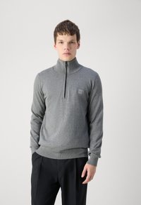 Pilkas zip-up megztinis su aukštu apykaklės ir rauktomis rankovėmis. Lygus mezgimo audinys su maža prekės ženklo etikete ant krūtinės. Juodos kelnės.