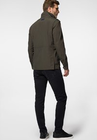 Jasnooliwkowa, lekka kurtka z wysokim kołnierzem, widocznymi szwami i bocznymi kieszeniami, w połączeniu z czarnymi dżinsami slim-fit i czarnymi butami.