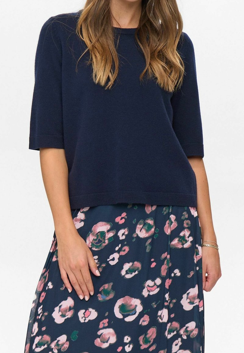 Femme portant un pull à manches courtes bleu marine et une jupe bleu marine ornée de motifs floraux roses et verts, la main posée sur la jupe.