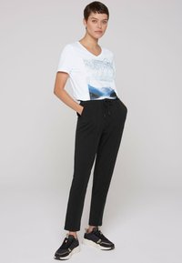 Witte t-shirt met bergafbeelding en tekst, gecombineerd met zwarte broek met koord en zwarte sneakers voor een casual uitstraling.