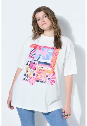 Jonge vrouw met golvend haar die een oversized wit T-shirt draagt met kleurrijke tropische en geometrische patronen, gecombineerd met een blauwe spijkerbroek.