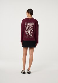 Bordeaux langemouwshirt met witte grafische afbeeldingen van voedselillustraties en tekst, gecombineerd met een zwarte corduroy rok en gemusterde schoenen.