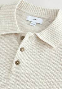 Beige strik polo shirt med ribbet krave, fire brune knapper og et struktureret stofmønster. Etiketten viser "NEXT" og størrelse "M."