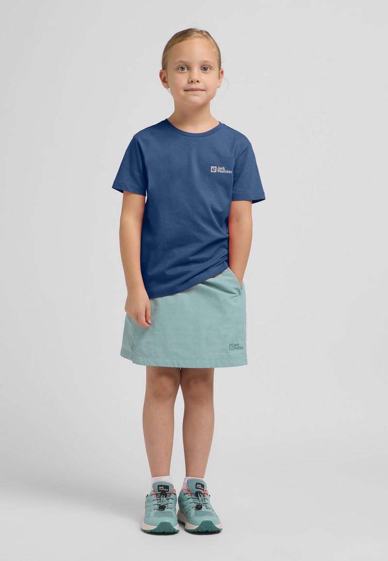 Marineblauw t-shirt met logo, lichtgroene rok en turquoise sportschoenen met zwarte en roze accenten, staand op een neutrale achtergrond.
