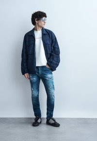 Άνδρας με σγουρά μαλλιά που φοράει ασημένια γυαλιά ηλίου, ναυτικό μπλε μπουφάν, λευκό πουκάμισο, σκισμένο μπλε τζιν και μαύρα αθλητικά παπούτσια, στέκεται μπροστά σε έναν απλό τοίχο.