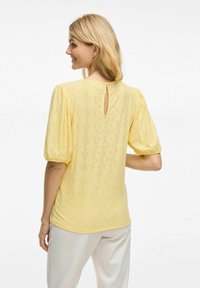 Blouse en coton jaune avec de courtes manches ballon, présentant un dos en forme de trou de serrure et un motif œillet décoratif. Texture lisse avec une silhouette ajustée.