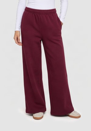 Personne portant un pantalon ample bordeaux avec une taille élastiquée, un haut blanc à manches longues et des baskets blanches sur un fond clair uni.
