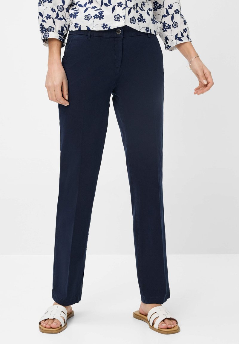 Marineblaue Baumwollhosen mit geradem Bein. Merkmale sind ein vorderer Knopfverschluss, Seitentaschen und abgekröpfter Saum. Kombiniert mit weißen Sandalen.