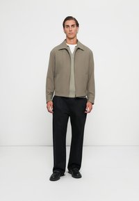Veste zippée vert olive clair, doublure en tricot, mannequin debout en pantalon et chaussures noirs, design minimaliste, texture lisse, sans motifs visibles.