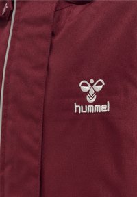 Bordeaux jas met een gladde textuur, voorzien van een witte geborduurde logo en een strakke ritssluiting, ontworpen voor casual gebruik.