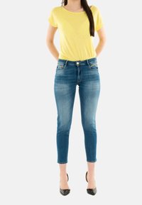 Camicia gialla a maniche corte abbinata a jeans skinny blu, caratterizzati da un classico design a cinque tasche e una leggera sfumatura, indossati con tacchi neri.
