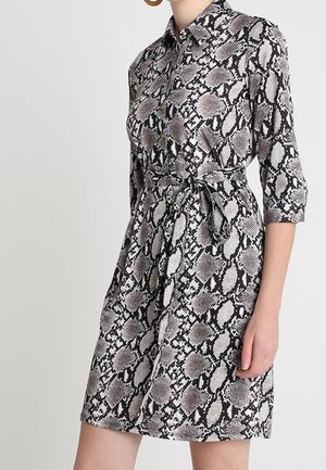 Femme portant une robe imprimé serpent, boutonnée, longueur genou, manches trois-quarts et ceinture nouée à la taille, debout devant un fond uni.