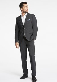 Donkergrijs pak met een getailleerde pasvorm, enkele knoopsluiting, notch-lapels, gecombineerd met een wit overhemd en een blauwe gepatterneerde pochet. Zwarte nette schoenen.