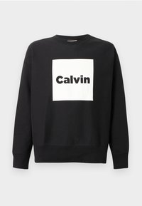 Čierna mikina s okrúhlym výstrihom a dlhými rukávmi, so stredovo umiestneným bielym štvorcom, na ktorom je slovo "Calvin" napísané tučnými čiernymi písmenami.