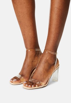 Sandales à talon transparent avec une base nude, présentant une sangle transparente sur les orteils et un talon bloc clair, une sangle à la cheville avec détail de boucle.