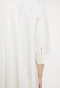 Bílá pruhovaná košile s rukávovým pletem a manžetami na knoflíky. Má dlouhé rukávy a rovný střih. Lehký, hladký textilní povrch.