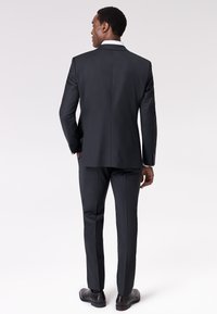 Completo scuro con una vestibilità su misura, mostrato da dietro. Presenta un'unica spacco, una texture liscia e scarpe scure coordinate.
