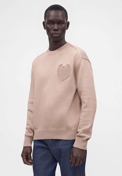 Homme portant un pull en maille beige avec un détail en forme de cœur texturé sur la poitrine et un jean bleu, debout devant un fond uni.