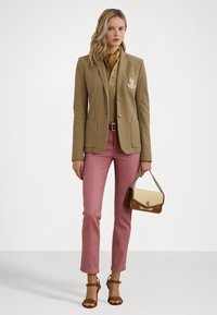 Blazer tan avec un logo brodé, superposé sur un polo beige. Associé à un pantalon rose ajusté et un sac à main bicolore. Talons ouverts marron.