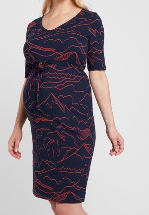 Zwangere vrouw in een marineblauwe zwangerschapsjurk met rode line-art van bergen en paarden, voorzien van een ceintuur in de taille en ellebooglange mouwen.