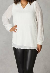 Donna che indossa una blusa bianca a maniche lunghe con texture e polsini arricciati, pantaloni in pelle nera e una collana d'oro, in posa con una mano sui fianchi.