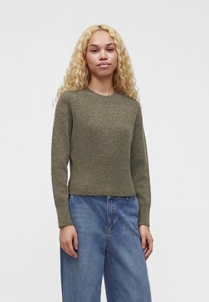 Jeune femme aux longs cheveux blonds bouclés portant un pull vert olive et un jean bleu ample, debout devant un fond uni.