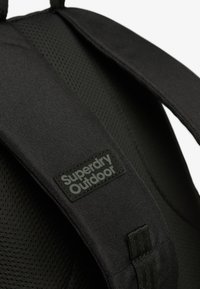 Zwarte stoffen rugzak met gewatteerde, gestructureerde banden en een "Superdry Outdoor" logo label. Materiaal heeft een gladde afwerking en een duurzame constructie.