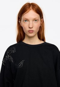 Sudadera negra con un corte holgado, adornada con rhinestones plateados dispuestos en un patrón gráfico en un lado.