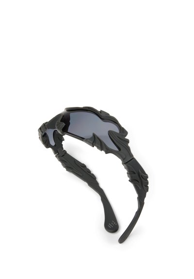 Zwarte zonnebril met hoekige, vlamachtige textuur aan de montuur en donker getinte glazen op een witte achtergrond.