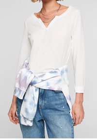 Vit långärmad topp med V-ringning, tillsammans med en pastellfärgad tie-dye-tröja knuten runt midjan och blå jeans.