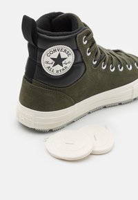 Olivově zelené vysoké tenisky vyrobené z semiše a kůže, s bílou gumovou podrážkou, stříbrnými očky a kulatou logovkou. Obsahuje tkaničky.