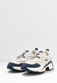 Sneaker bianche e blu navy con tomaia textured, sistema di allacciatura, linguetta imbottita e suola spessa con un design di battistrada prominente.