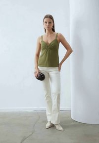 Olivgrünes geripptes Trägershirt mit einem vorderen Twist-Detail, kombiniert mit weißen Schlaghosen und einer schwarzen Clutch. Die Stilettos sind crèmefarben.