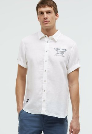 Mann trägt weißes kurzärmeliges Hemd mit Knopfleiste und dem Schriftzug „Camp David Surf“ sowie blaue Hose vor schlichtem hellem Hintergrund.