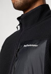Veste en polaire noire avec un col haut et fermeture éclair à l'avant. Comprend une poche poitrine en nylon noir contrastant avec un détail de logo.