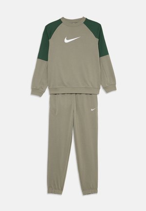 Olivgrüner Nike Trainingsanzug mit dunkelgrünen Schulterpanelen an dem Sweatshirt und weißen Nike-Logos auf der Brust und am Hosenbein.