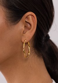 Goldene Ohrhänger mit einem gedrehten Design, polierter Oberfläche und glatter Textur, die von einem durchstochenen Ohr hängen.