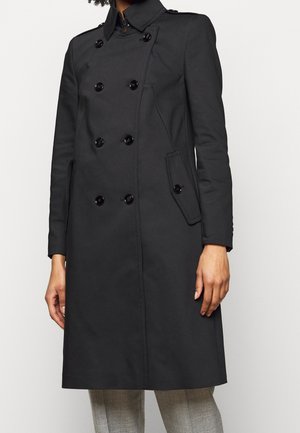 Black double-breasted trench coat with wide collar, buttoned flap pockets, and long sleeves worn over light gray pants.