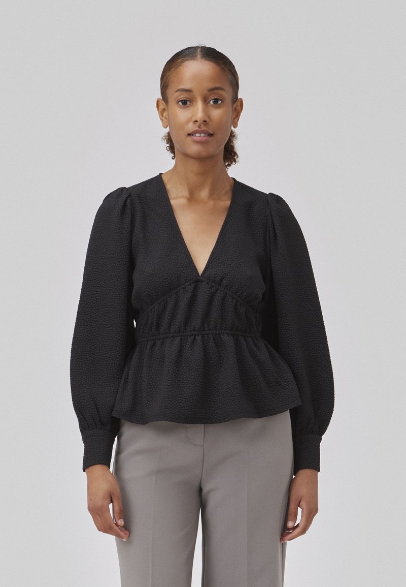 Black textured blouse with a deep V-neck, puffed sleeves, and gathered waistline, paired with light grey trousers.