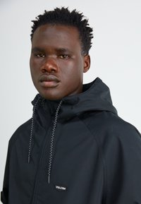 Chaqueta negra con capucha y cordones ajustables, hecha de una tela suave, que presenta un pequeño parche de logo blanco en el lado izquierdo del pecho.