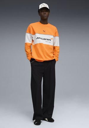 Homme portant un sweat-shirt McLaren Racing orange et blanc, un pantalon noir, des chaussures Puma noires et une casquette Puma blanche, debout les mains dans les poches.