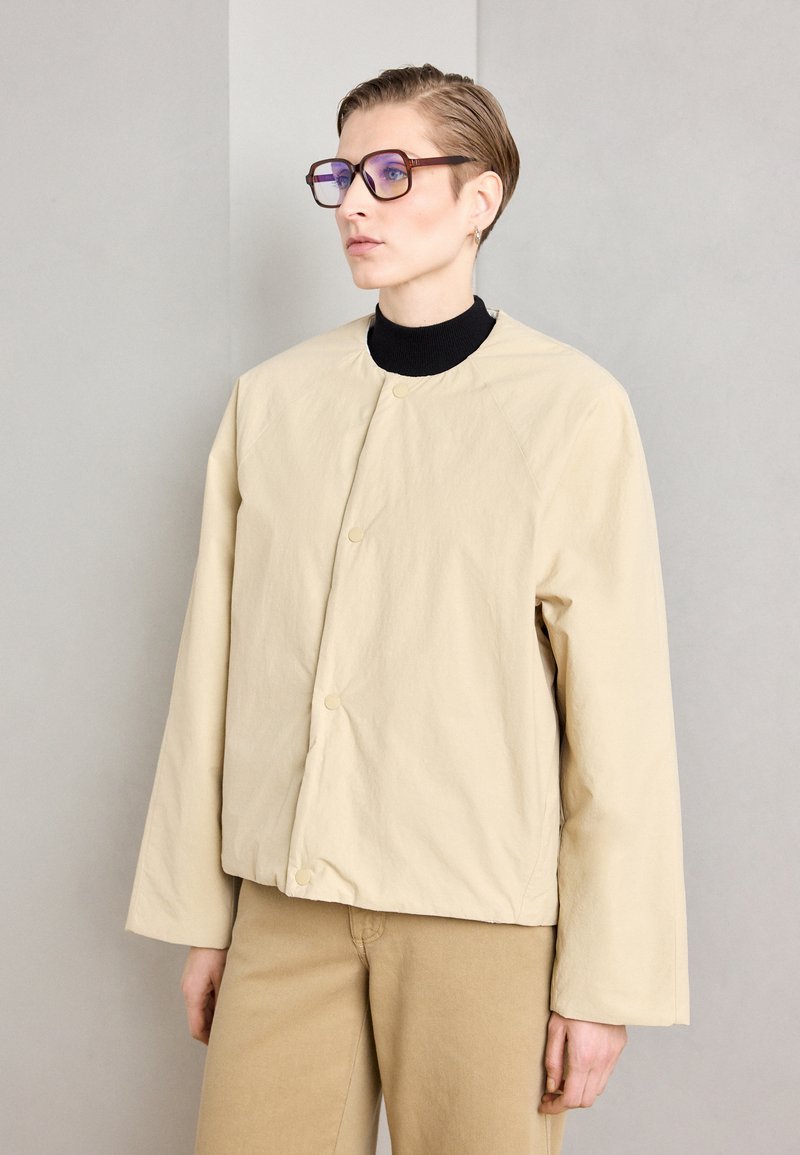 Beige, oversized jacket with a rounded neckline, raglan sleeves, and snap closures. Worn over a black turtleneck and light beige pants.